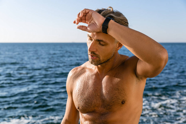 уставший взрослый мужчина без рубашки вытирает пот со лба после тренировки на берегу моря
 - Фото, изображение