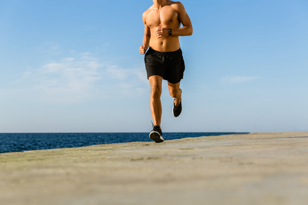 cropped shot of shirtless sportsman jogging on seashore - Photo, Image