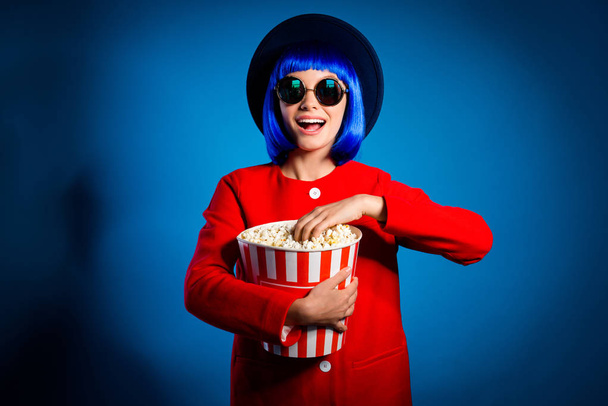 Portrait of excited laughing girl in eyewear red coat headwear holding big bucket with pop corn looking at camera isolated on blue background. Entertainment free time concept - Fotó, kép