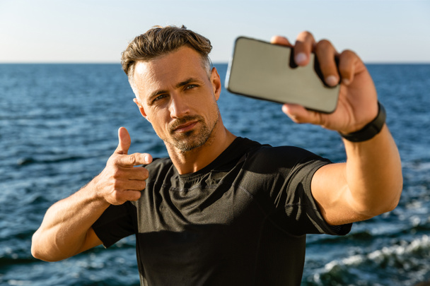 όμορφος ενήλικος άνδρας λαμβάνοντας αυτοπορτρέτα με smartphone και δείχνοντας στην κάμερα στην παραλία - Φωτογραφία, εικόνα