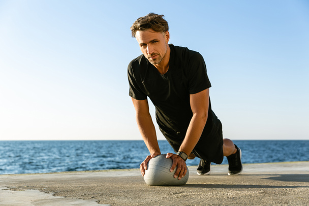 apuesto deportista adulto haciendo flexiones con la bola en forma en la orilla del mar y mirando a la cámara
 - Foto, imagen