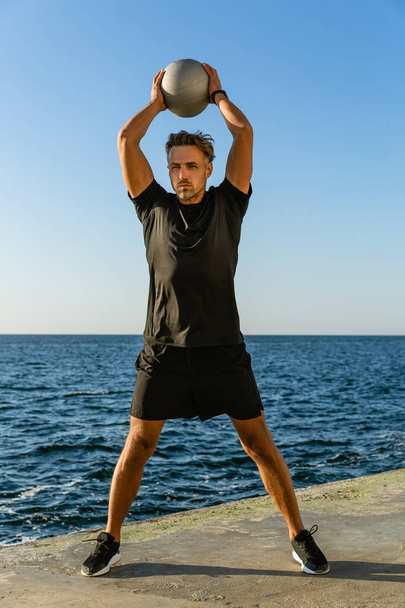 apuesto deportista adulto haciendo ejercicio con la bola en forma y sosteniéndolo por encima de la cabeza en la orilla del mar
 - Foto, imagen