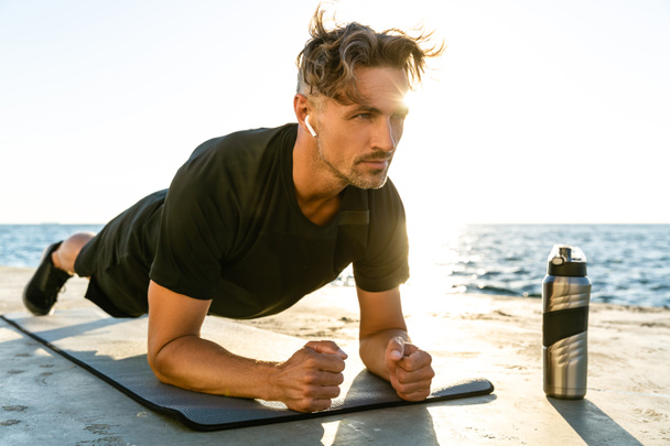 handsome adult sportsman with wireless earphones doing plank exercise on seashore - Photo, Image