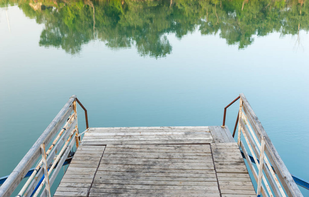 Old wooden and rusted metal jetty on a lake or river with still water and reflections of the surrounding green trees - Φωτογραφία, εικόνα