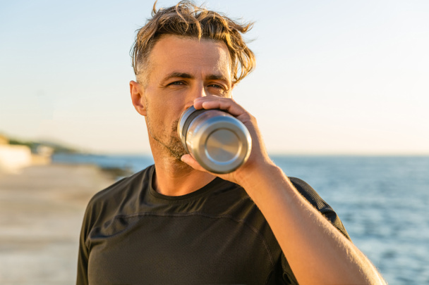 handsome adult man drinking water from fitness bottle on seashore in front of sunrise after training - Foto, imagen