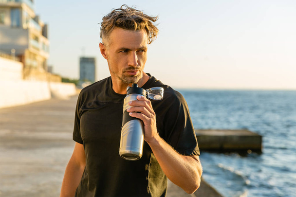 handsome adult sportsman with grey hair holding fitness bottle on seashore in front of sunrise looking away - Foto, imagen