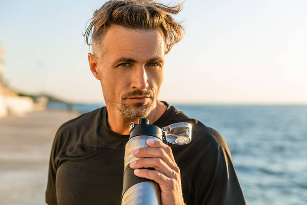 красивий дорослий чоловік з фітнес-пляшкою на березі моря перед сходом сонця
 - Фото, зображення