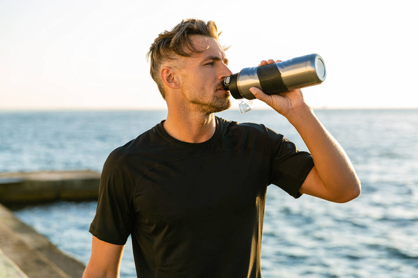 adult sportsman drinking water from fitness bottle on seashore in front of sunrise - Photo, Image