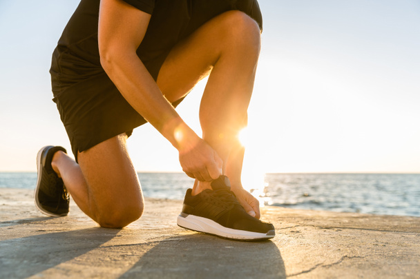 recortado tiro de deportista atando zapatillas de deporte en la orilla del mar delante de la salida del sol
 - Foto, Imagen