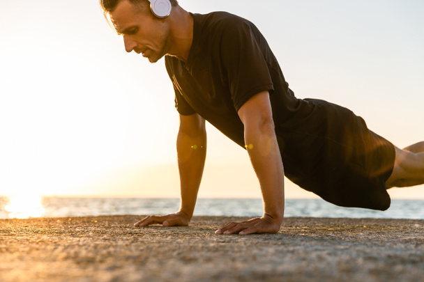 athletic adult man in headphones doing push ups on seashore - Photo, Image