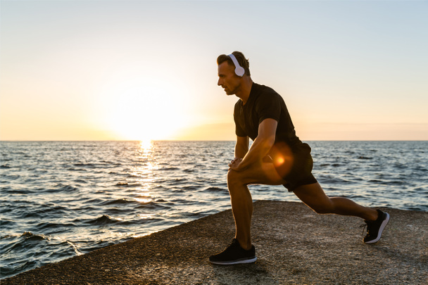 athletic adult man in headphones doing one legged squats during training on seashore - Photo, Image