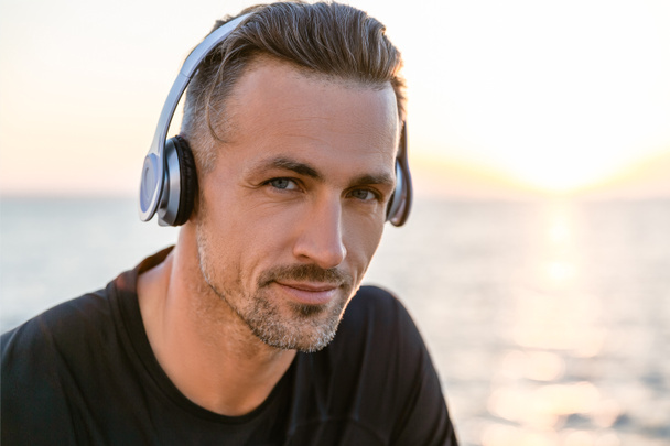close-up portrait of attractive adult man in wireless headphones looking at camera on seashore - Photo, Image