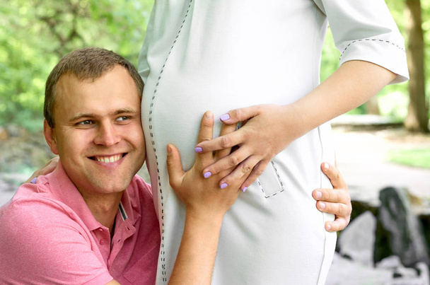 Un bel homme écoute le ventre de sa belle femme enceinte et sourit. Le mari écoute le ventre de sa femme. Joyeux famille enceinte drôle
. - Photo, image
