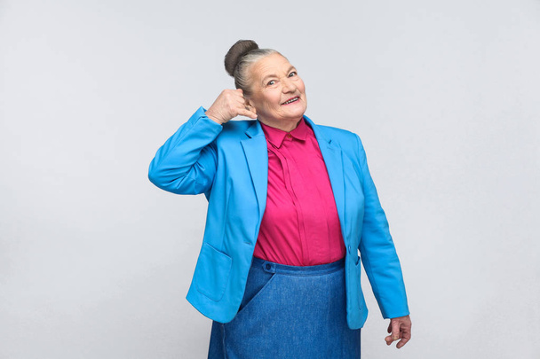 expressive grandmother with bun hairstyle in blue suit and pink shirt showing call me sign and smiling on gray background - Foto, immagini