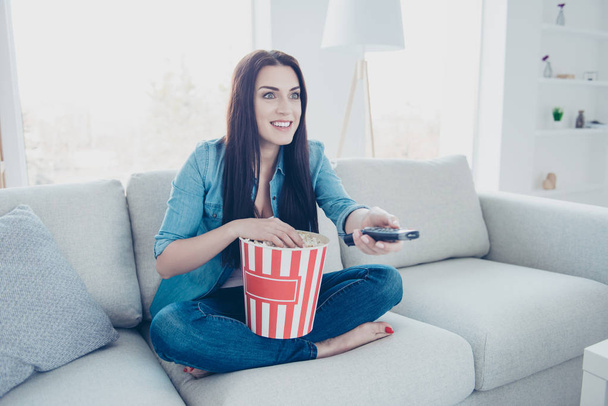 Portrait of cheerful excited woman sitting on sofa in modern white room with crossed legs having bucket with pop-corn using remote controller searching favorite channel - Foto, Bild