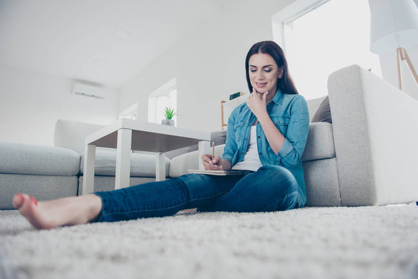 Low angle view of thoughtful pensive woman sitting on carpet near sofa in modern white apartment holding hand on chin writing list to do in notepad using pen wearing jeans outfit - Fotografie, Obrázek