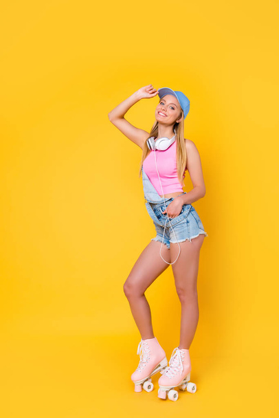 Full body, size portrait of slim fit sporty active girl in denim overall with headset on neck holding hand in pocket of shorts looking at camera isolated on bright background - Foto, Imagem