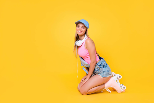 Portret van funky positieve meisje zittend op de knieën dragen rolschaatsen denim outfit met hoofdtelefoon op nek stralendste glimlach kijken camera geïsoleerd op gele achtergrond - Foto, afbeelding
