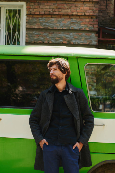 handsome man in grey coat leaning back on green car near house  - Foto, Imagen