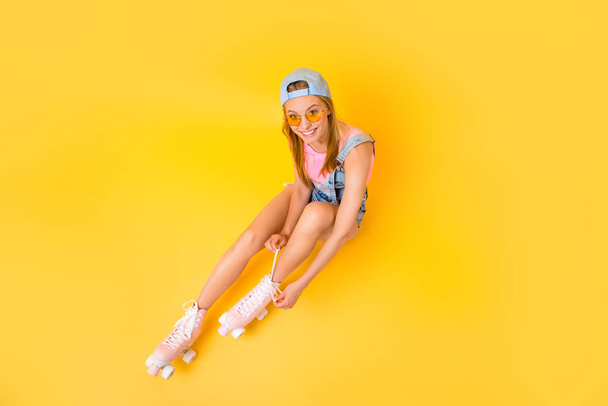 Top view portrait of cheerful joyful chick putting on roller skates tying shoelaces looking at camera isolated on yellow background sitting on floor ground, street outdoor urban lifestyle concept - Foto, Imagem