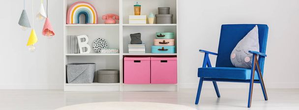 Panorama of a cute pillow on a blue armchair and a bookcase with boxes and decorations in a fun child bedroom interior with white walls - Φωτογραφία, εικόνα