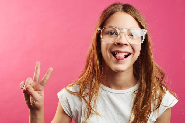 Portrait of brown-haired girl in glasses showing tongue out and peace symbol on pink background - Foto, Imagem