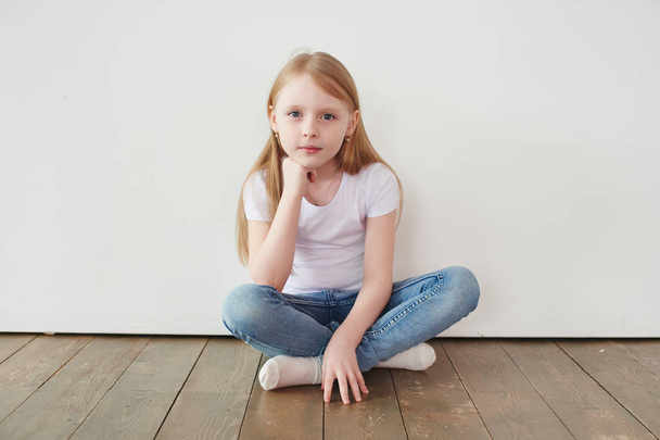 Portrait of little blonde girl in white t-shirt toothy smiling and looking at camera while sitting on  wooden floor near white wall - Photo, Image
