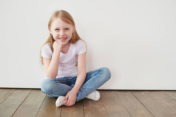 Portret mała Blondynka w biały t-shirt toothy, uśmiechając się i patrząc na kamery, siedząc na drewnianej podłodze w pobliżu biała ściana - Zdjęcie, obraz