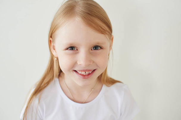 Portrait of little blonde girl in white t-shirt toothy smiling and looking at camera on white studio background, close-up - Foto, Imagem
