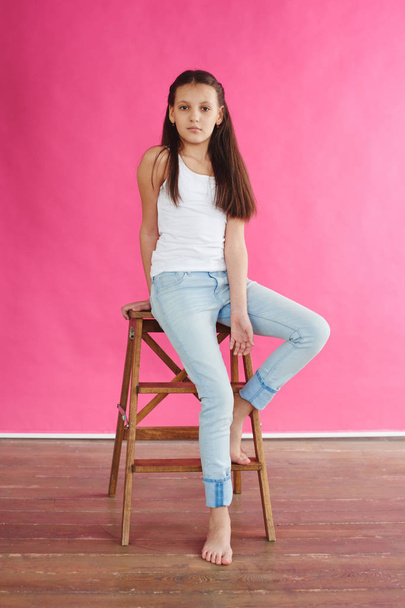 girl model in white tank top and jeans sitting on wooden chair and posing on pink background  - Photo, image