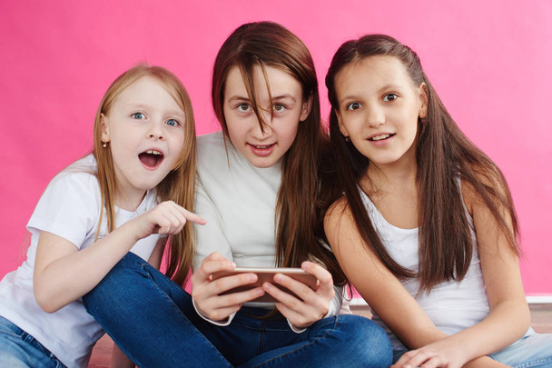 cute girls wearing casual clothes playing games on smartphone while sitting on pink studio background - Фото, изображение