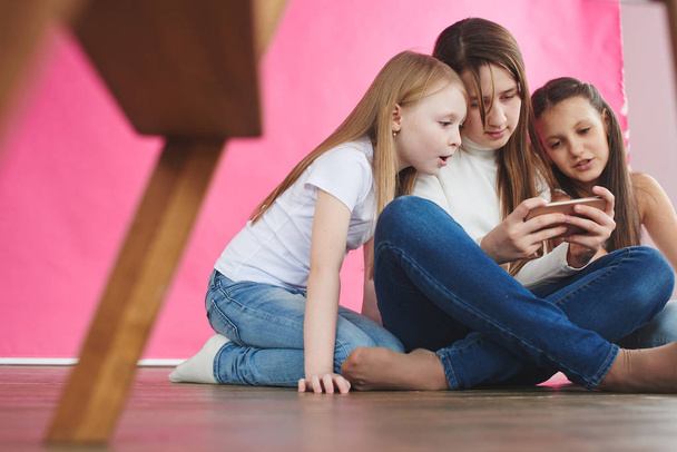 cute girls wearing casual clothes playing games on smartphone while sitting on pink studio background - Foto, afbeelding