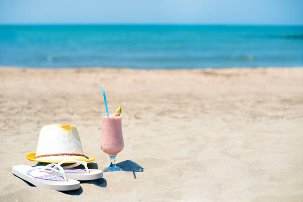 Strawberry fresh smoothie juice, hat and slippers near sea on sandy beach - Photo, Image
