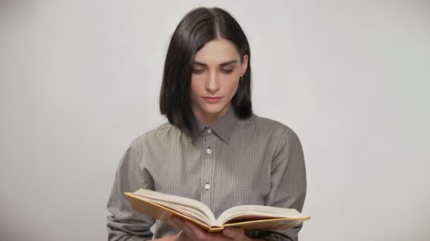Young pretty woman with short brown hair holding book and reading, then looking in camera and smiling, white background - Video