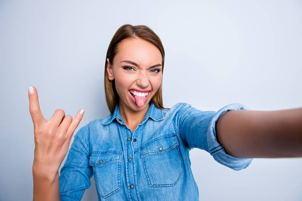 Self portrait of cool crazy girl shooting selfie on front camera showing rock and roll symbol with tongue out isolated on grey background - Φωτογραφία, εικόνα