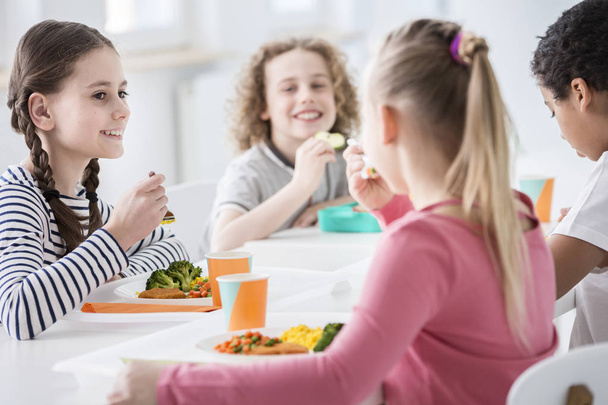 Smiling girl eating vegetables during lunch break with friends at school - Foto, imagen