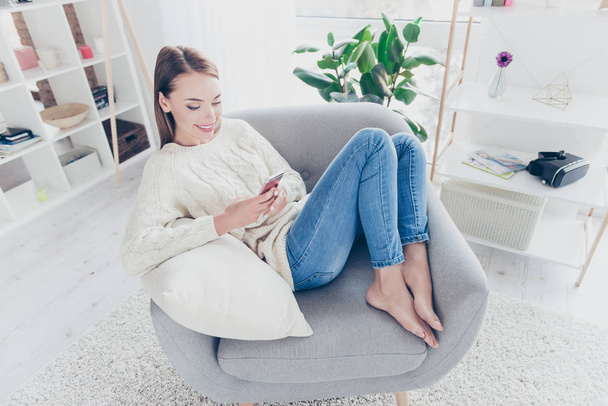 High angle view of cute addicted blogger in jeans sweater sitting on chair in modern living room with interior having smart phone in hands using wi-fi 3G internet - Fotoğraf, Görsel