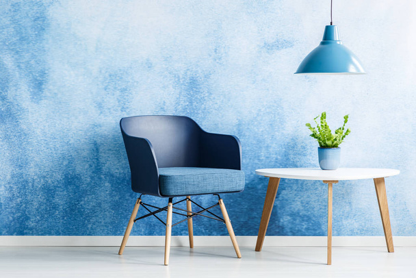 Lamp above white table with plant next to navy blue armchair in simple living room interior - Foto, Bild