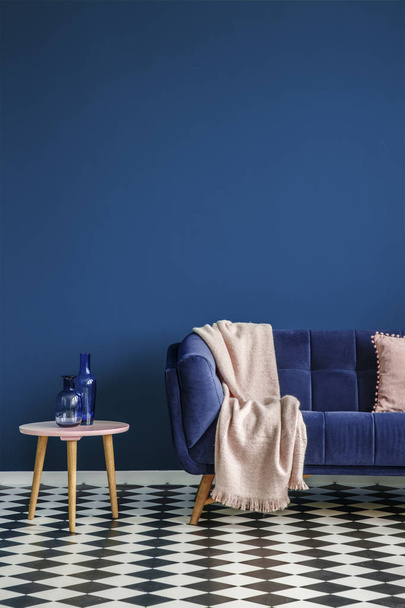 Chessboard floor, sofa with a blanket and stool with glass decorations on an empty navy blue wall in living room interior. Real photo - Photo, image