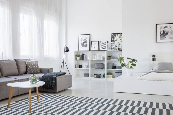 Table on patterned carpet in open space interior with white bed and grey corner sofa. Real photo - Foto, Imagem
