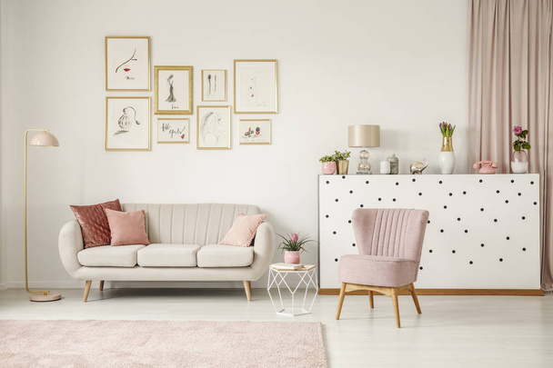 Pink chair next to table and sofa in living room interior with posters and gold lamp - Fotoğraf, Görsel