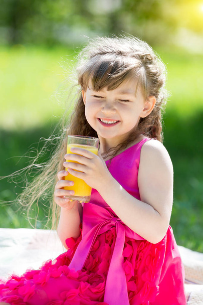 Little girl in red dress on picnic in park sits and drinks orange juice. - Фото, изображение