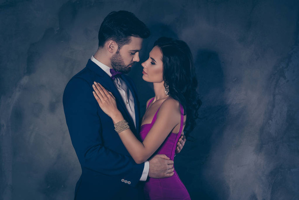 Side view portrait of attractive lovely couple, virile harsh gentleman in tuxedo embracing his pretty charming lady on waist  looking at each other face to face isolated on grey background - Φωτογραφία, εικόνα