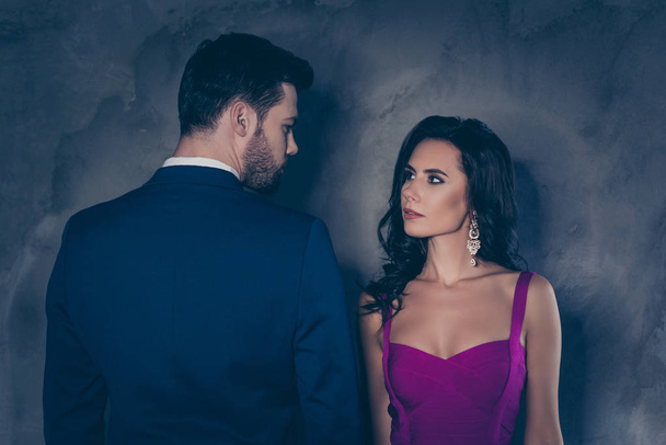 Rear view portrait of brunet gentleman in tuxedo and pretty lady with front view in purple outfit with jewelry earring curls decollete, couple looking at each other isolated on grey background - Fotografie, Obrázek