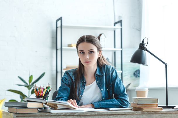 atractiva chica adolescente sentada en el escritorio y mirando a la cámara mientras estudia en casa
 - Foto, Imagen