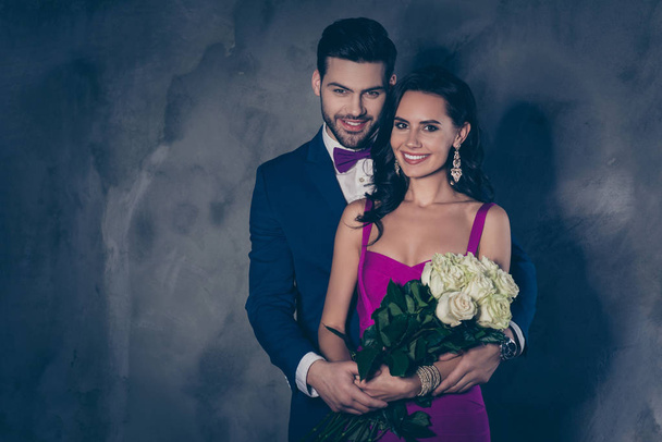 Portrait of pretty lady in purple dress with jewelry accessories hispanic business rich gentleman in tuxedo with bowtie holding white roses in hands, lovely romantic spouses isolated grey background - Fotoğraf, Görsel