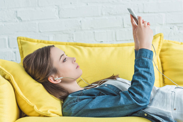 side view of attractive girl in earphones lying on sofa and listening music with smartphone - Fotografie, Obrázek