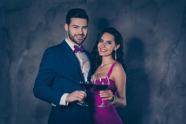 Portrait of positive couple having wineglasses with red wine in hands looking at camera, gentleman in tuxedo with bow charming hispanic latin lady earring in purple outfit isolated on grey background - Photo, image