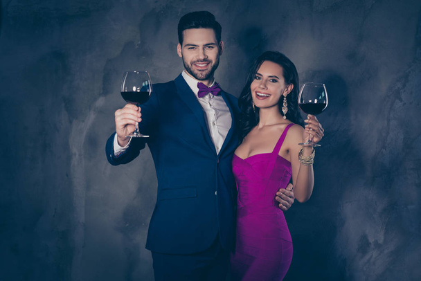 Portrait of cheerful festive couple having wineglasses with red wine in raised hands looking at camera, gentleman in tux with bowtie charming lady in purple outfit isolated on grey background - Фото, изображение