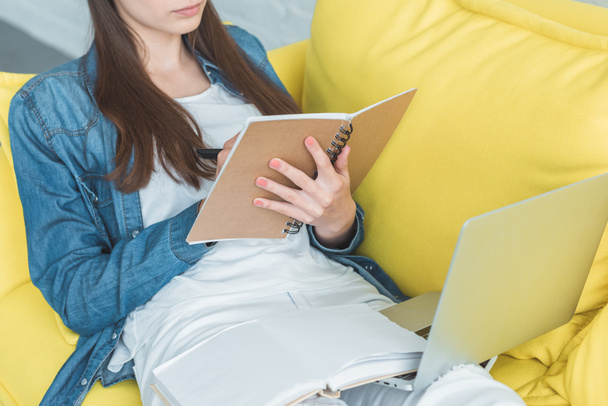 cropped shot of girl writing in notebook and using laptop on sofa at home - Foto, imagen